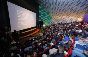 Uma foto tirada da parte superior do auditório, mostrando a grandiosidade do evento. O foco principal é o grande telão branco que exibe um texto jurídico em português. O auditório está cheio, evidenciando as poltronas vermelhas dispostas em patamares e o detalhe decorativo na parede lateral em quadrados verdes e brancos.