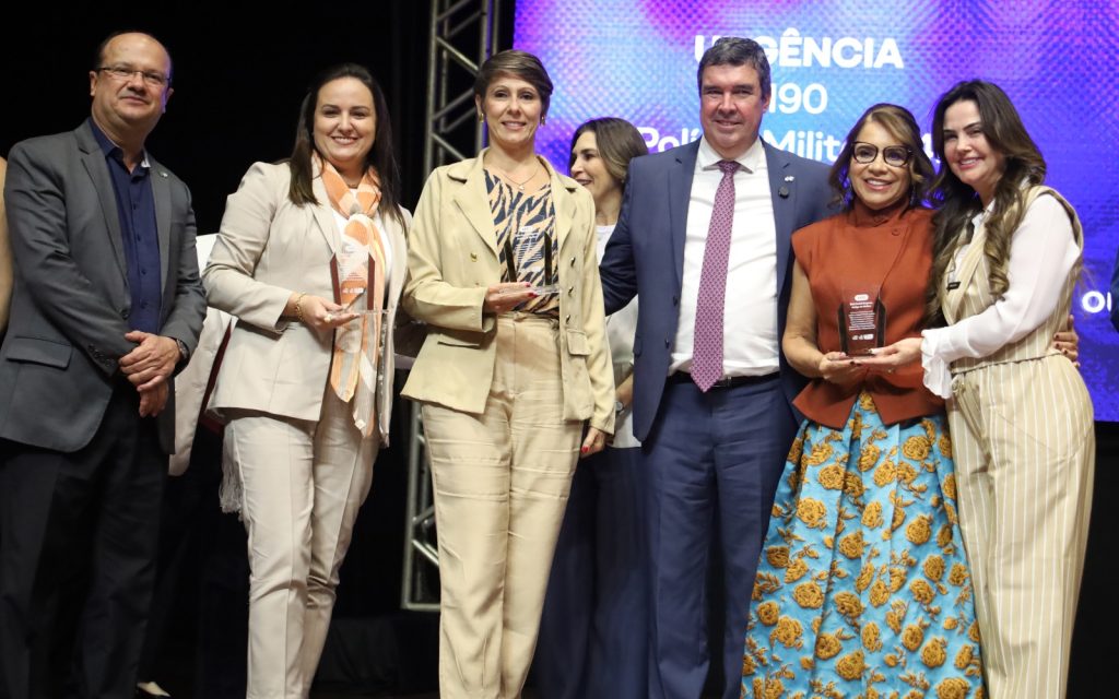 Fotografia de um grupo de seis pessoas (quatro mulheres e dois homens) posando para uma foto oficial em um palco. Três das mulheres seguram troféus de acrílico transparente. No centro, um homem de terno azul e gravata bordô sorri para a câmera. À direita, uma mulher usa uma saia longa azul com bordados florais amarelos e um colete laranja, segurando seu prêmio ao lado de outra mulher em um conjunto listrado bege. Ao fundo, uma tela de LED exibe informações de segurança pública, incluindo o número 190 e a frase "Polícia Militar".