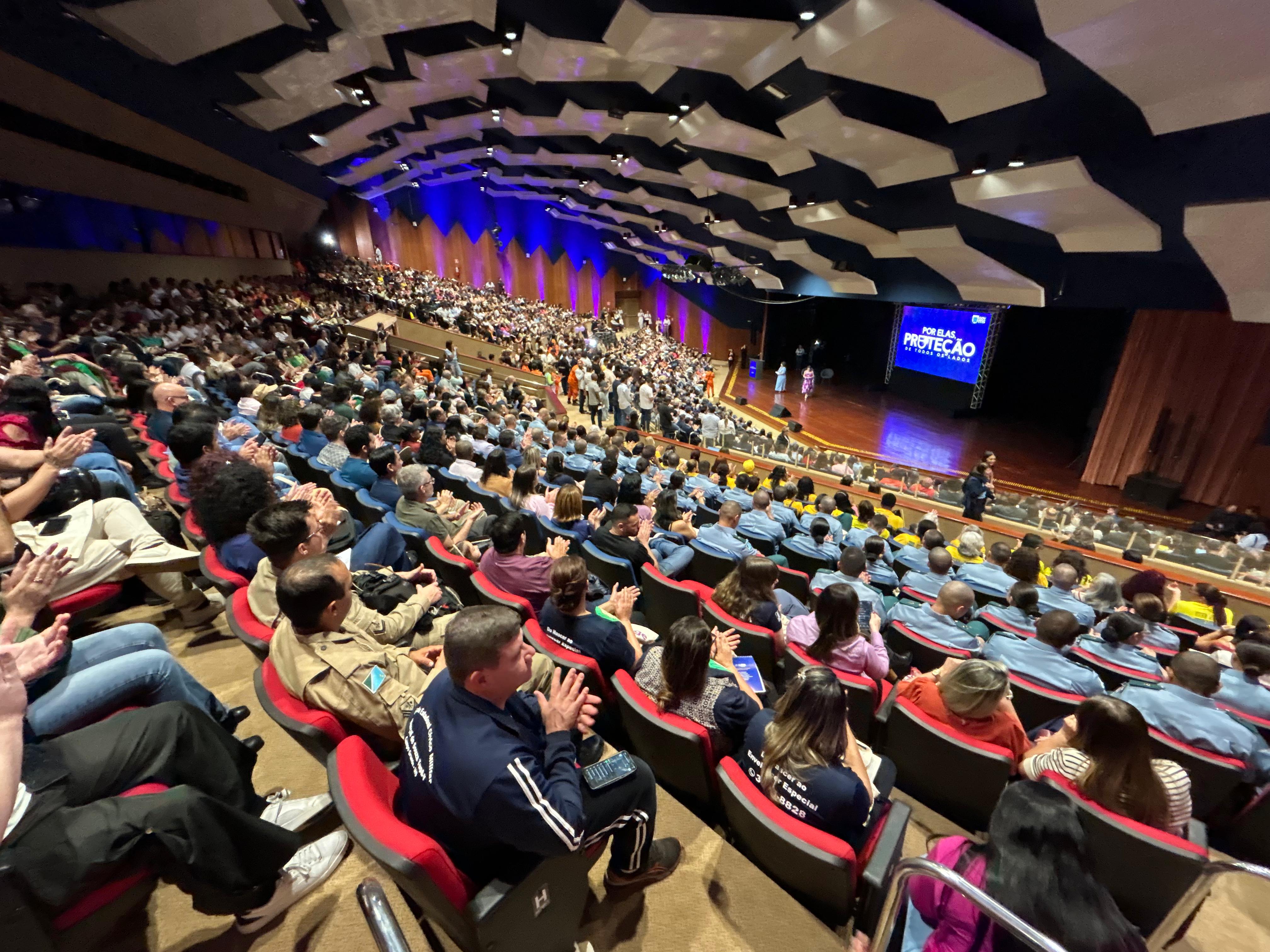 Fotografia em ângulo aberto de um grande auditório lotado. Centenas de pessoas estão sentadas em poltronas vermelhas, muitas delas vestindo uniformes de forças de segurança (camisas azuis e cáqui). Ao fundo, no palco, há um telão azul com o título do evento: "POR ELAS, PROTEÇÃO DE TODOS OS LADOS". O teto do auditório possui painéis acústicos brancos em formatos geométricos e a iluminação lateral é em tons de azul e roxo. O público parece aplaudir ou observar o palco atentamente.