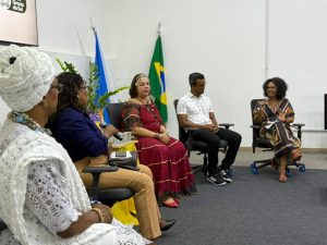 Visão lateral de uma mesa de debate. Cinco pessoas estão sentadas em cadeiras de escritório pretas dispostas em semicírculo. Em destaque, no lado esquerdo, uma mulher negra com vestes tradicionais brancas e turbante. Ao lado dela, uma mulher de blusa azul fala ao microfone. No centro, uma mulher com trajes tradicionais ciganos em tons de vermelho e dourado. Ao fundo, as bandeiras do Brasil e do Mato Grosso do Sul.