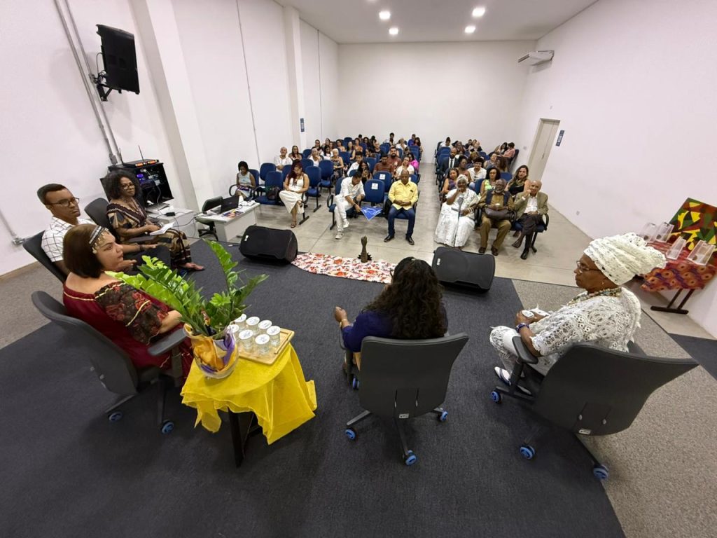 Uma foto tirada de um ângulo alto e amplo, mostrando o palco e a plateia. No palco, os palestrantes estão sentados em semicírculo. À frente deles, no chão, há um trilho de tecido estampado. A plateia ocupa cadeiras azuis e é composta por um grupo diverso de pessoas que acompanham o evento atentamente. As paredes do auditório são brancas e o teto possui iluminação embutida.