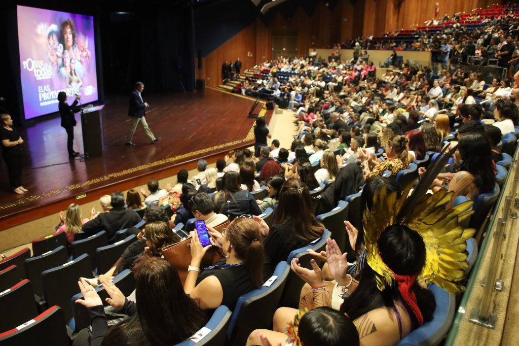 Pra todos verem: Vista da plateia lateral do teatro. Em destaque no primeiro plano, as costas de uma mulher indígena usando um grande cocar de penas amarelas. O público ao redor aplaude a apresentação que ocorre no palco ao fundo.
