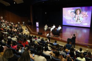 Pra todos verem: Fotografia panorâmica tirada da parte superior da plateia de um teatro lotado. O público, majoritariamente feminino, observa o palco onde há um enorme telão com a frase "Elas, Protagonistas". No palco, uma pessoa fala ao microfone em um púlpito iluminado.