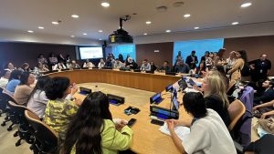 Fotografia em plano aberto de uma sala de conferências nas Nações Unidas. Diversas pessoas estão sentadas ao redor de uma grande mesa semicircular de madeira clara. No centro da mesa, há placas de identificação azuis, incluindo uma onde se lê "RED CEA". Ao fundo, uma tela de projeção exibe um texto. Nas laterais e ao fundo, outras pessoas acompanham a reunião em pé ou sentadas em cadeiras extras. O ambiente tem iluminação suave e teto com diversas luminárias embutidas.