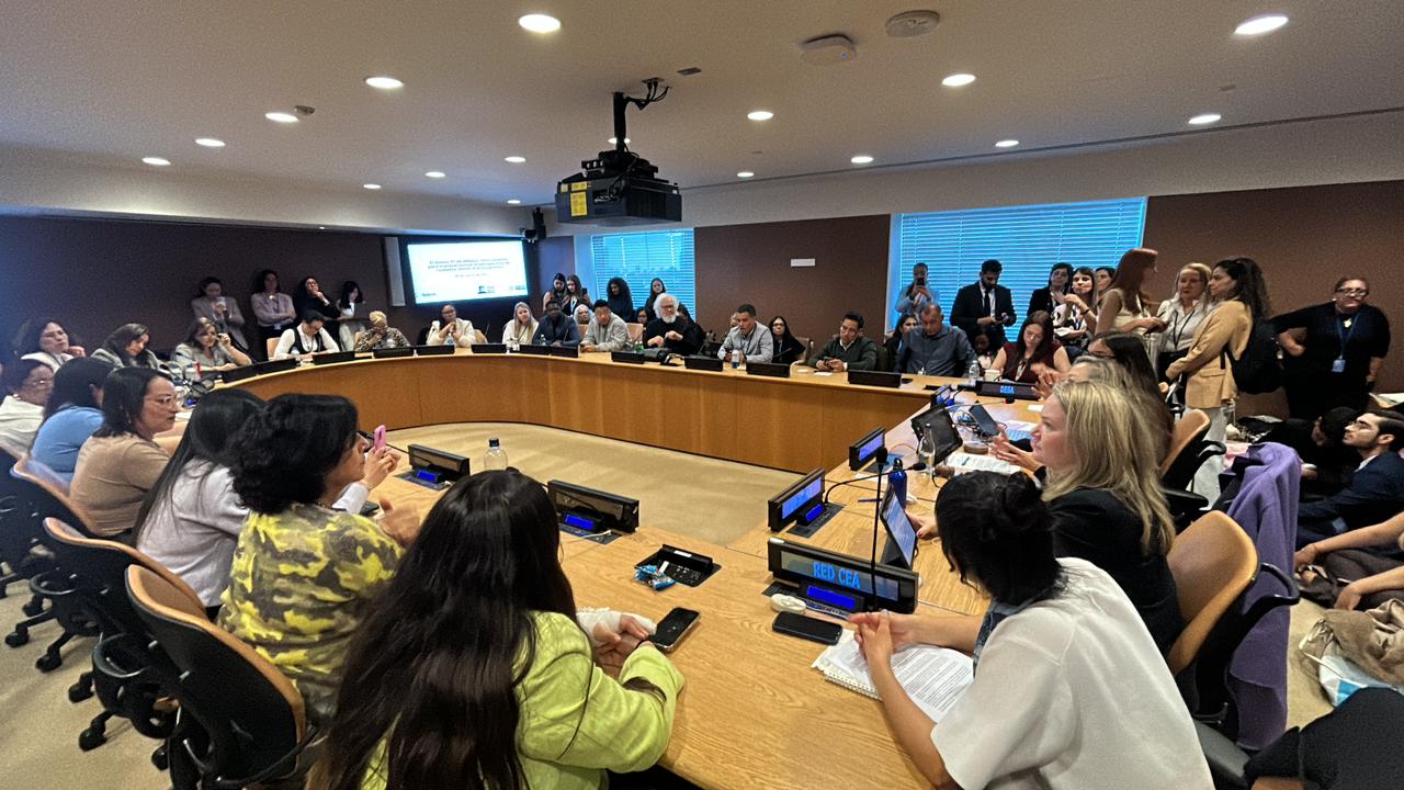 Fotografia em plano aberto de uma sala de conferências nas Nações Unidas. Diversas pessoas estão sentadas ao redor de uma grande mesa semicircular de madeira clara. No centro da mesa, há placas de identificação azuis, incluindo uma onde se lê "RED CEA". Ao fundo, uma tela de projeção exibe um texto. Nas laterais e ao fundo, outras pessoas acompanham a reunião em pé ou sentadas em cadeiras extras. O ambiente tem iluminação suave e teto com diversas luminárias embutidas.