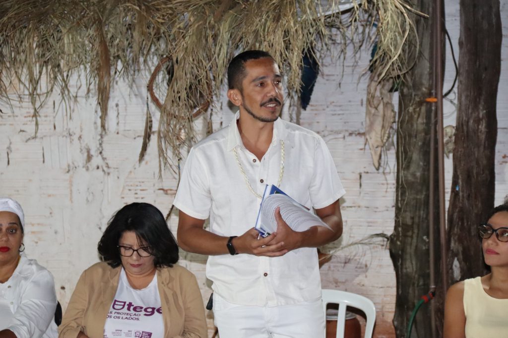 Um homem jovem, de pele parda e cabelos curtos, está de pé falando ao público. Ele veste uma camisa social branca e segura papéis nas mãos. Ele está em um ambiente que remete a um terreiro, com teto de palha seca ao fundo e paredes de tijolo aparente pintadas de branco. Ao lado dele, sentadas, estão duas mulheres participando da conversa. O clima é de diálogo e atenção.