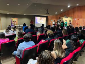 Fotografia do interior de um auditório com luz suave. A visão é de trás para a frente, mostrando a plateia composta majoritariamente por mulheres sentadas em poltronas vermelhas. No palco, à esquerda, há um banner vertical com a frase "Todas são todas importantes". Ao centro, uma mulher de blusa vermelha fala ao microfone em frente a uma tela de projeção que exibe um cartaz colorido. À direita do palco, um grupo de seis mulheres está de pé, observando a apresentação. As paredes do auditório têm acabamento em madeira e tons neutros.