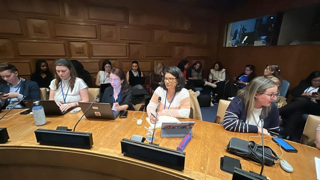 Fotografia de ângulo médio mostrando uma bancada de madeira curva em uma sala de reuniões com paredes revestidas de painéis de madeira esculpida. Manuela está sentada ao centro, usando uma blusa listrada de branco e rosa claro, escrevendo em um caderno. Ao lado dela, outras mulheres operam laptops. À frente de cada assento, há microfones de base preta e equipamentos de tradução ou votação. No canto superior direito, vê-se uma janela de vidro que dá para uma cabine de tradução simultânea.