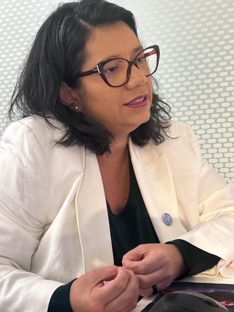 Retrato em plano médio de uma mulher (Manuela) de pele clara e cabelos escuros ondulados na altura dos ombros. Ela usa óculos de armação escura, batom cor de vinho e um blazer branco sobre uma blusa escura. Na lapela do blazer, há um pequeno bóton azul com o emblema da ONU. Ela está levemente voltada para a direita, com as mãos unidas à frente, parecendo falar durante uma reunião. O fundo é uma parede branca com textura de pequenos relevos ovais.