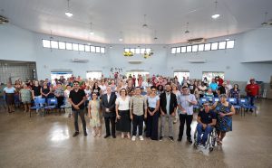 Foto panorâmica em um grande salão circular com teto alto e paredes cinza-claras. No centro do primeiro plano, um grupo de cerca de dez autoridades e organizadores (homens e mulheres) posa lado a lado. Atrás deles, dezenas de idosos e participantes do evento estão de pé, ocupando o centro do salão. Ao fundo, janelas altas e retangulares circundam a parte superior da parede. O ambiente é amplo e bem iluminado.