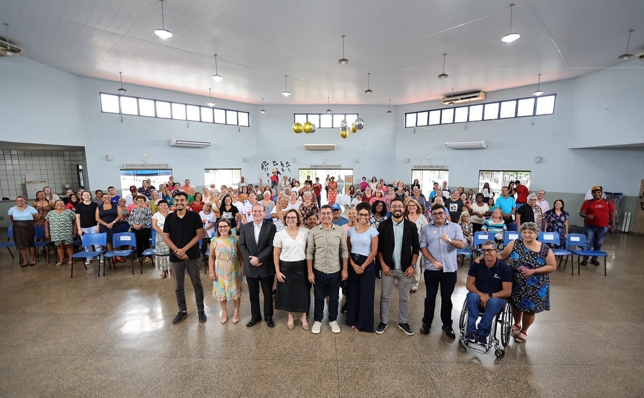 Foto panorâmica em um grande salão circular com teto alto e paredes cinza-claras. No centro do primeiro plano, um grupo de cerca de dez autoridades e organizadores (homens e mulheres) posa lado a lado. Atrás deles, dezenas de idosos e participantes do evento estão de pé, ocupando o centro do salão. Ao fundo, janelas altas e retangulares circundam a parte superior da parede. O ambiente é amplo e bem iluminado.