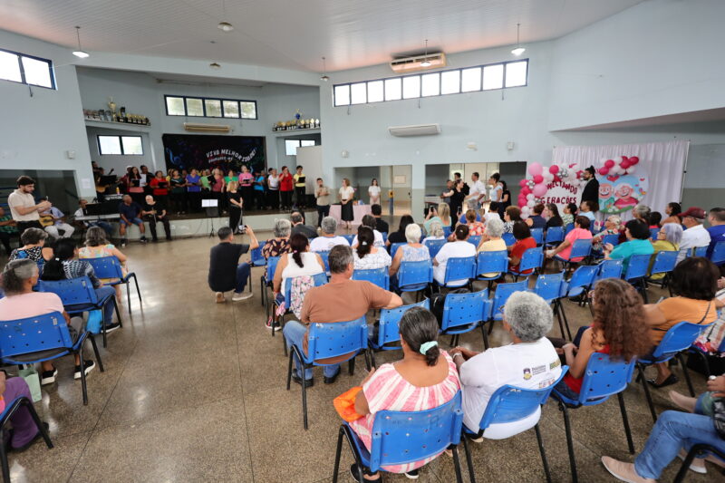 Vista do fundo do salão mostrando o público sentado em cadeiras de metal azul, voltado para um palco ao fundo. No palco, um coral de idosos se apresenta. À direita, há um arco de balões rosa e brancos com um painel onde se lê "Conectando Gerações". O chão é de granilite polido e reflete a iluminação do teto.
