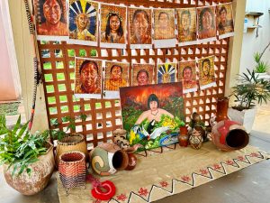 Exposição cultural em um ambiente externo. Painéis com retratos de pessoas indígenas estão pendurados em uma parede de cobogós. No chão, sobre um tapete artesanal, há cestarias, vasos de cerâmica decorados e uma tela pintada com uma figura indígena cercada por animais da fauna brasileira.