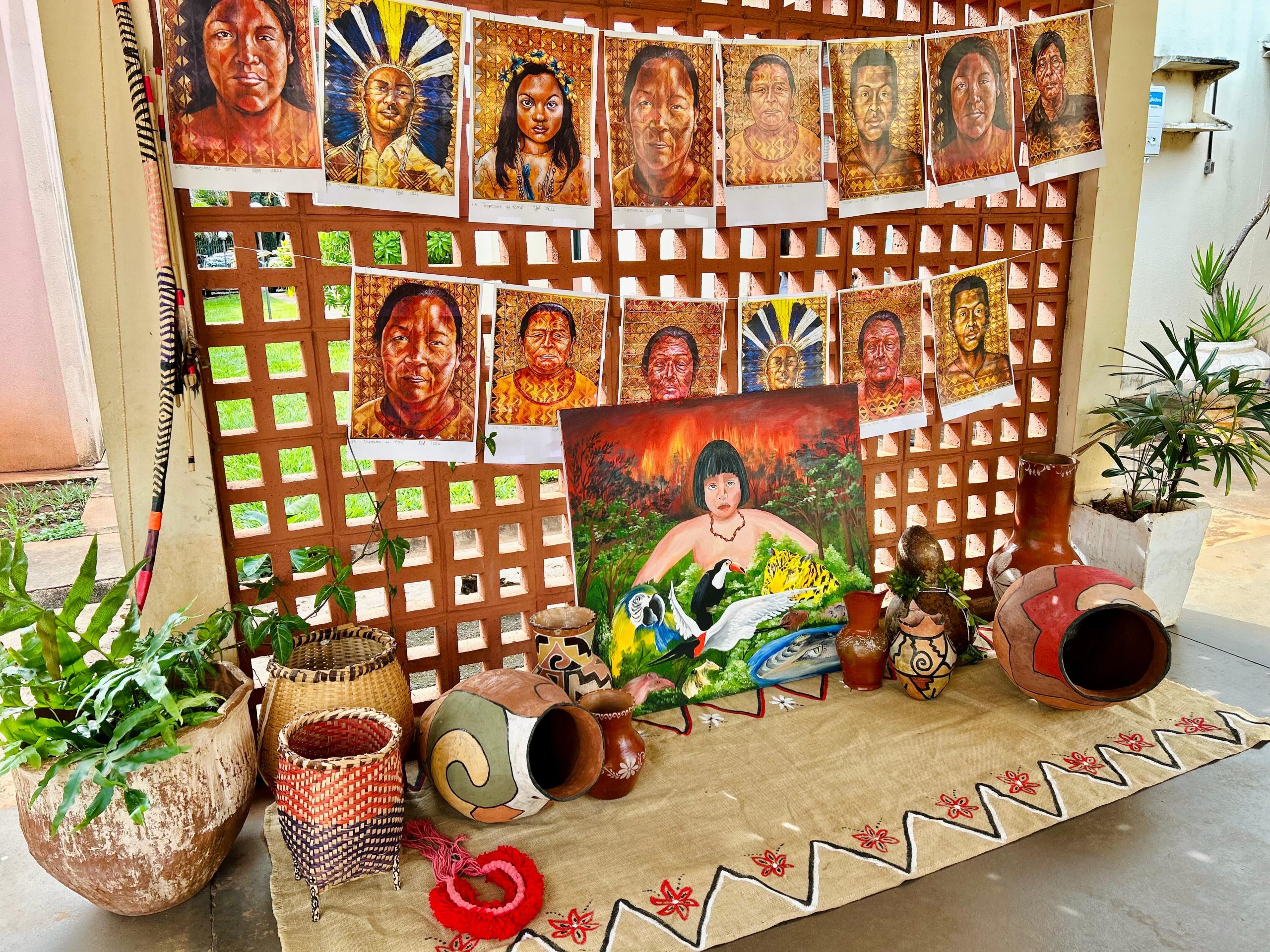 Exposição cultural em um ambiente externo. Painéis com retratos de pessoas indígenas estão pendurados em uma parede de cobogós. No chão, sobre um tapete artesanal, há cestarias, vasos de cerâmica decorados e uma tela pintada com uma figura indígena cercada por animais da fauna brasileira.