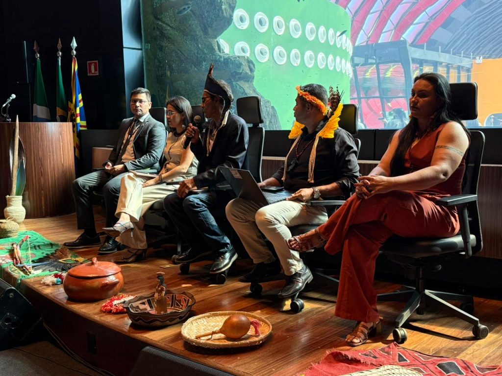 Mesa redonda composta por cinco pessoas em um auditório. Entre os participantes, homens e mulheres indígenas com adornos tradicionais dividem espaço com pessoas em trajes sociais. No chão, à frente da mesa, há uma exposição de objetos culturais como maracás, cerâmicas e cestarias. Ao fundo, vê-se um grande aquário.