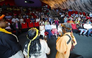 Fotografia de plano médio capturada de um ângulo elevado em um auditório moderno. No centro, três pessoas indígenas de costas para a câmera conversam com um grupo de autoridades sentadas na primeira fila. O homem indígena à direita veste uma camisa alaranjada, cocar de sementes e segura um microfone. O auditório está cheio, com poltronas vermelhas e uma parede curva revestida com painéis de madeira em tons de cinza e bege.