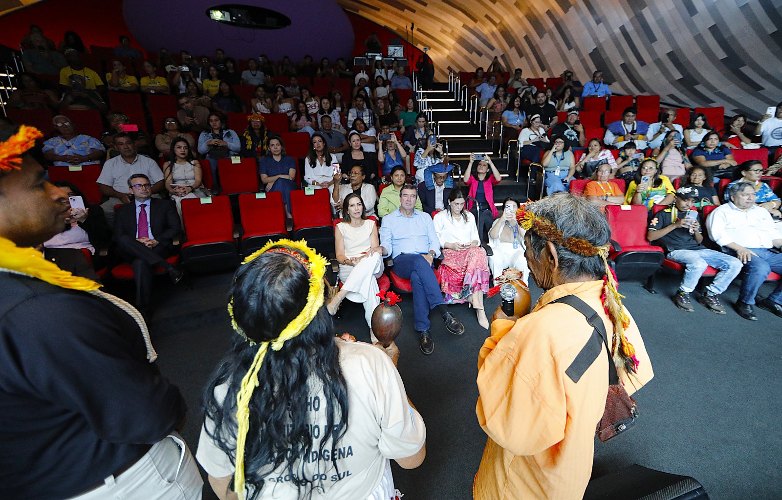 Fotografia de plano médio capturada de um ângulo elevado em um auditório moderno. No centro, três pessoas indígenas de costas para a câmera conversam com um grupo de autoridades sentadas na primeira fila. O homem indígena à direita veste uma camisa alaranjada, cocar de sementes e segura um microfone. O auditório está cheio, com poltronas vermelhas e uma parede curva revestida com painéis de madeira em tons de cinza e bege.