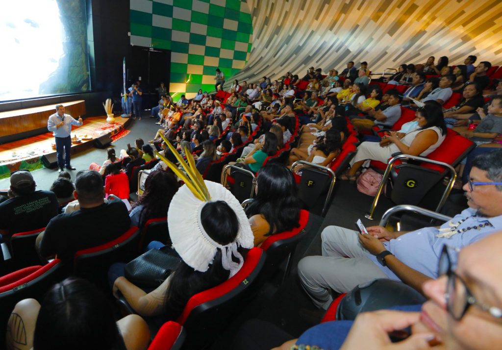 Vista panorâmica do auditório a partir da lateral da plateia. À esquerda, em um pequeno palco, um homem de camisa azul clara fala ao microfone. A plateia é diversa, composta por pessoas indígenas e não indígenas. Uma mulher indígena em destaque na parte inferior, de costas, usa um grande cocar de penas brancas e amarelas. O teto e a parede lateral são curvos, com um padrão geométrico de quadrados verdes e brancos ao fundo.