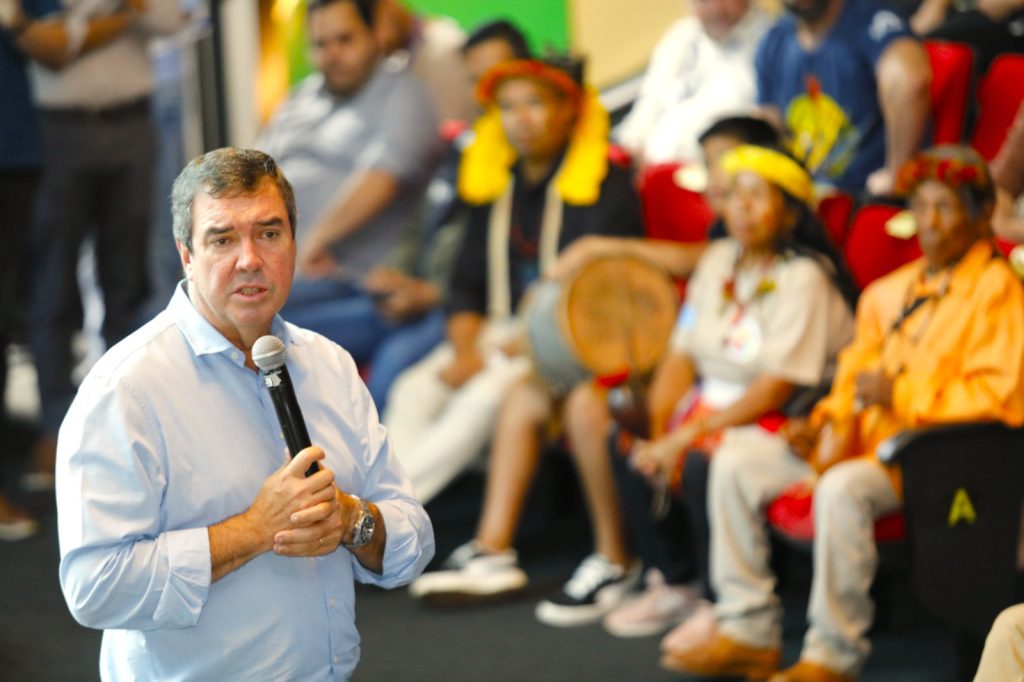 Close-up de um homem de pele clara, cabelos curtos e grisalhos, vestindo uma camisa social azul clara. Ele segura um microfone com as duas mãos à altura do peito e fala com expressão atenta. O fundo está desfocado, mas é possível ver pessoas indígenas sentadas em poltronas vermelhas, usando adornos tradicionais amarelos e vermelhos.