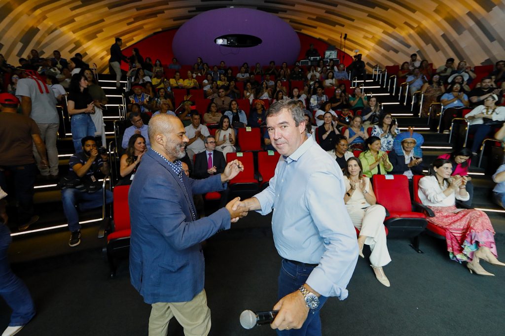 Fotografia em ângulo levemente superior mostrando o interior de um auditório moderno com teto curvado e amadeirado. Em primeiro plano, dois homens se cumprimentam com um aperto de mãos e sorrisos. O homem à esquerda veste um blazer azul sobre calça bege; o homem à direita veste uma camisa social azul clara, calça jeans e segura um microfone. Ao fundo, as poltronas vermelhas do auditório estão ocupadas por um público diverso, incluindo pessoas com trajes casuais e algumas com adereços indígenas (cocares). A iluminação foca no palco e nas primeiras fileiras, criando um clima de evento oficial ou conferência.