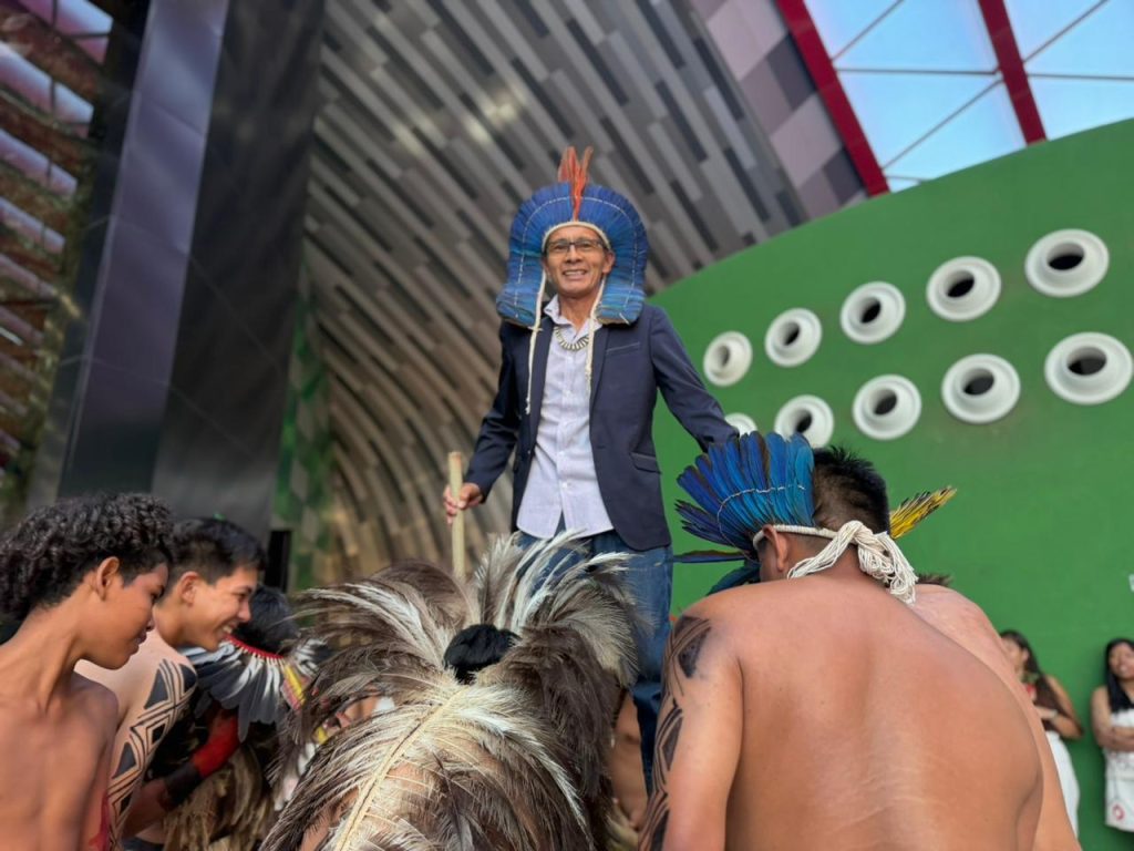 Um homem indígena sorridente está em pé em um nível elevado. Ele usa um cocar de penas azuis e laranjas, óculos, camisa clara, paletó azul-marinho e calça jeans. À sua frente, em primeiro plano e levemente desfocados, outros homens indígenas estão de costas, sem camisa, com pinturas corporais pretas e grandes adornos de penas nas costas.