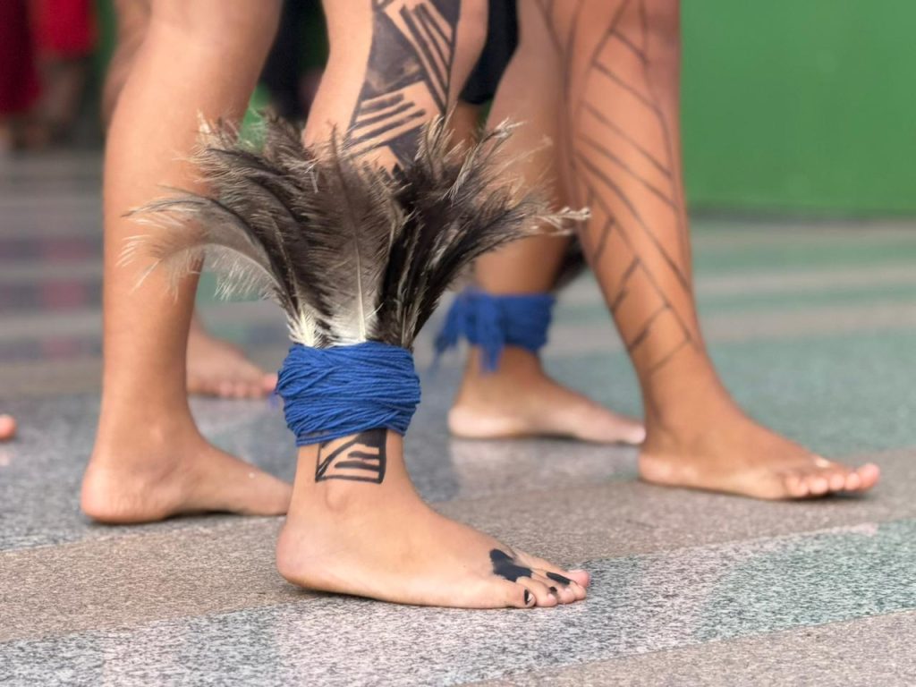 Close-up focado nos pés e pernas de jovens indígenas. Eles estão descalços sobre um chão de granito claro. Uma das pernas apresenta pinturas geométricas pretas e um adorno de penas escuras preso ao tornozelo por cordas de lã azul vibrante. Outro pé ao lado tem uma pequena pintura preta nos dedos.