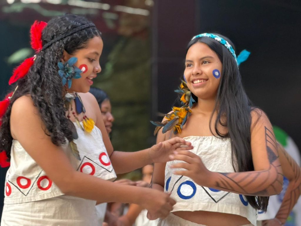 Duas jovens indígenas sorriem uma para a outra. Elas usam vestimentas claras com grafismos geométricos em vermelho e azul, pinturas faciais circulares e adornos de penas nos cabelos e orelhas.