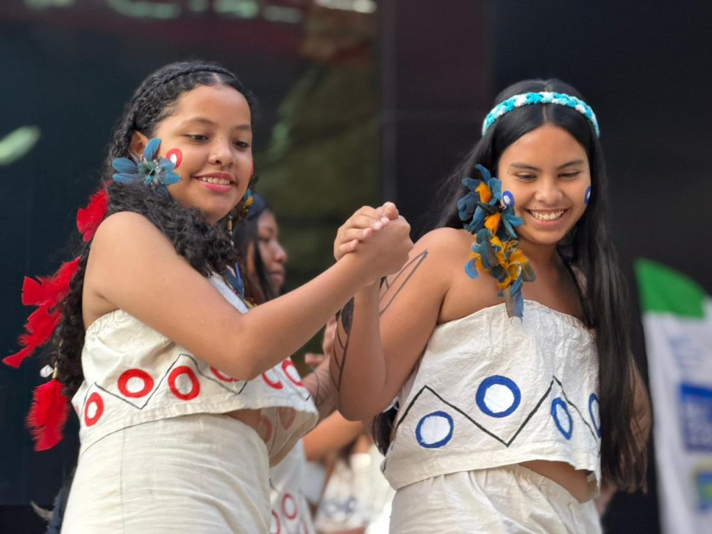 Close-up de duas jovens indígenas sorrindo e segurando as mãos uma da outra durante uma dança. Elas vestem trajes de tecido branco com desenhos geométricos circulares em vermelho e azul. Ambas têm flores azuis e penas coloridas adornando os cabelos e pinturas faciais vermelhas e azuis nas bochechas. O fundo está desfocado.