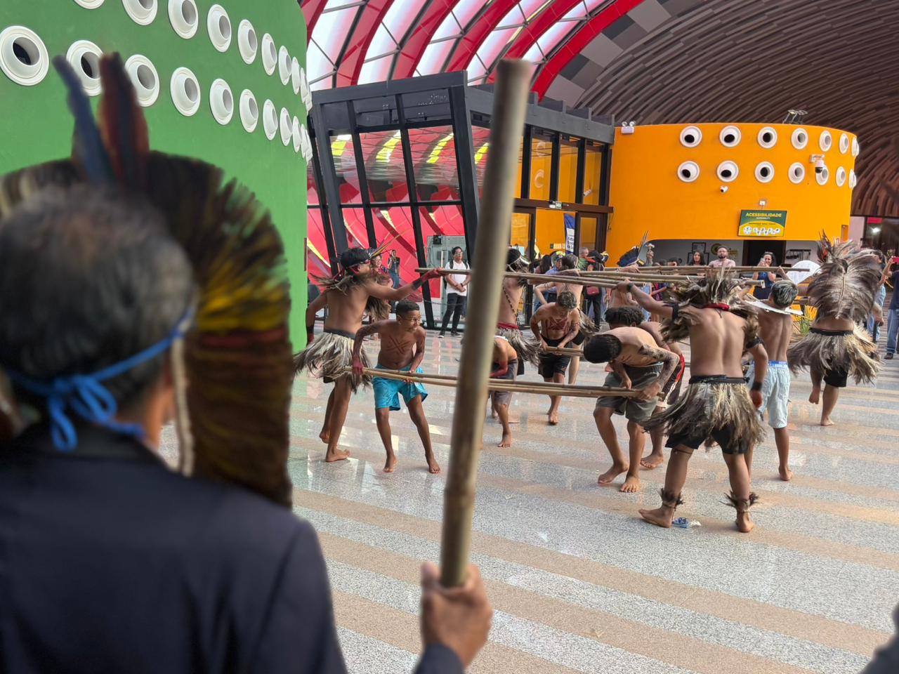 Jovens indígenas realizam uma apresentação cultural no Bioparque Pantanal. Eles estão em movimento, segurando bastões de bambu sobre o piso espelhado. Ao fundo, a arquitetura moderna do local exibe tons de verde e laranja. Em primeiro plano, a silhueta de uma pessoa com cocar observa a cena.