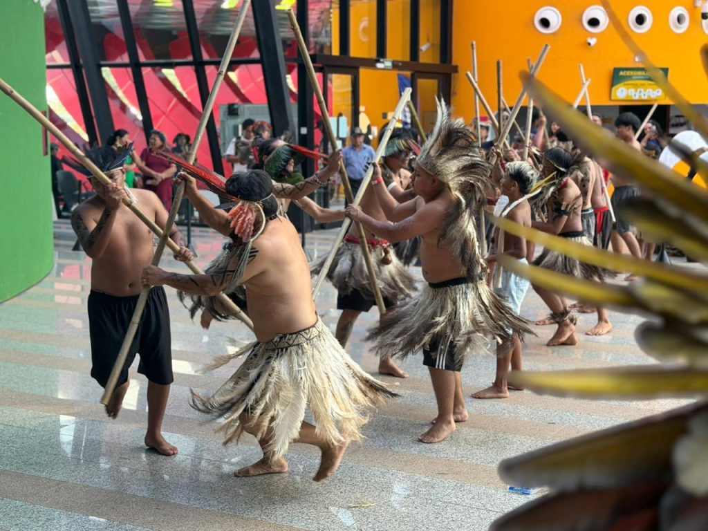 Fotografia de ação mostrando um grupo de homens indígenas realizando uma dança ou celebração em um espaço amplo e iluminado. Eles estão sem camisa, usam saias de fibras ou penas claras e carregam longas varas de madeira. Estão em movimento, com os corpos inclinados. Ao fundo, uma parede laranja vibrante com janelas circulares e uma estrutura de vidro.