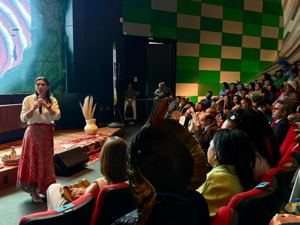 Uma mulher de pele clara, cabelos castanhos longos, vestindo uma blusa branca e uma saia longa vermelha estampada, fala ao microfone no palco do auditório. Ela está de perfil para a plateia. Em primeiro plano, vemos as cabeças de pessoas na plateia, incluindo um homem indígena com um cocar de penas escuras. Ao fundo, um grande telão exibe uma imagem azulada.