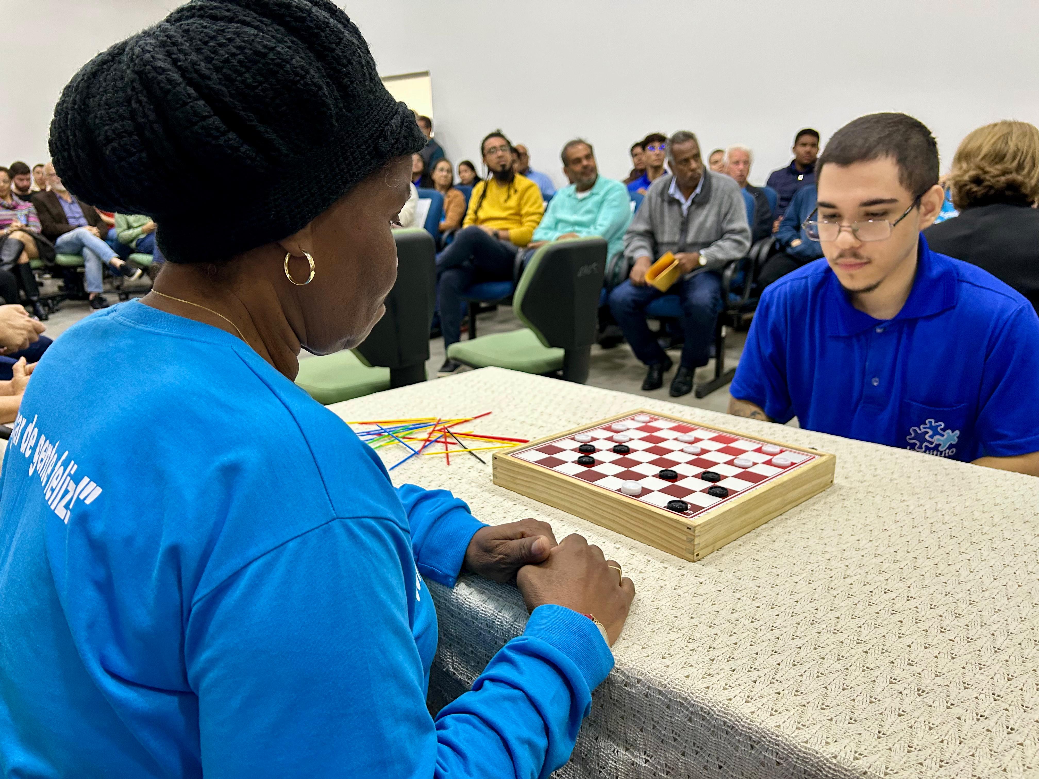 Fotografia em plano médio de uma interação entre gerações. Em primeiro plano, de costas para a câmera, uma mulher negra usa uma touca de crochê preta e uma camiseta azul de manga longa. Ela está sentada à mesa em frente a um jovem de pele clara, que usa óculos e camiseta polo azul. Entre eles, há um tabuleiro de damas de madeira sobre uma toalha bege rendada. O jovem observa o tabuleiro com atenção. Ao lado, há palitos coloridos do jogo "Pega Varetas". Ao fundo, várias pessoas de diferentes idades estão sentadas em cadeiras de auditório, observando a cena em um ambiente de paredes brancas.