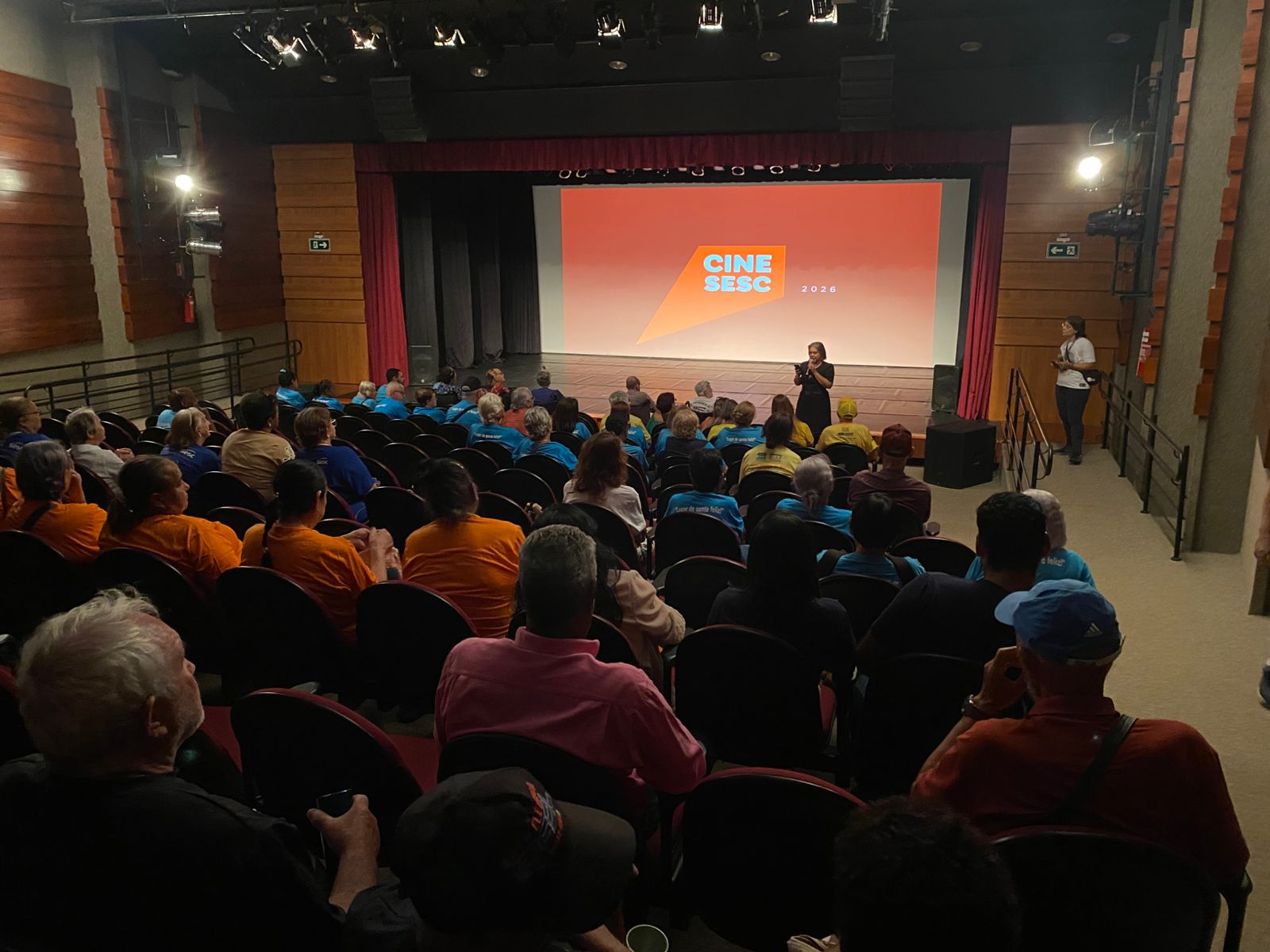 Fotografia horizontal em plano aberto tirada de trás de uma plateia em um teatro ou cinema. No palco, ao centro, uma mulher de preto fala ao microfone diante de um telão laranja vibrante onde se lê "CINE SESC 2026". A plateia é composta majoritariamente por idosos, muitos vestindo camisetas coloridas em tons de azul, laranja e amarelo. O ambiente tem luz suave, paredes revestidas de madeira e poltronas escuras. No canto direito, outra pessoa da equipe observa o evento em pé.