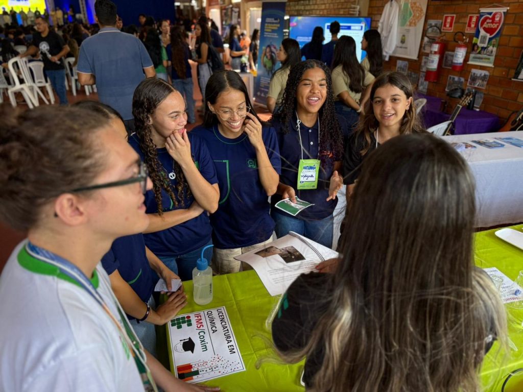 Estudantes em volta de uma bancada com o letreiro "Licenciatura em Química - IFMS/Coxim". Eles sorriem e conversam enquanto observam materiais educativos e um frasco sobre a mesa coberta com toalha verde.