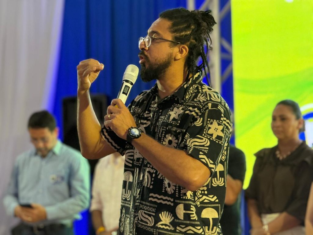 Um homem negro com dreads presos, barba e óculos fala ao microfone. Ele veste uma camisa preta com estampas geométricas brancas e gesticula com a mão direita durante sua fala no palco.