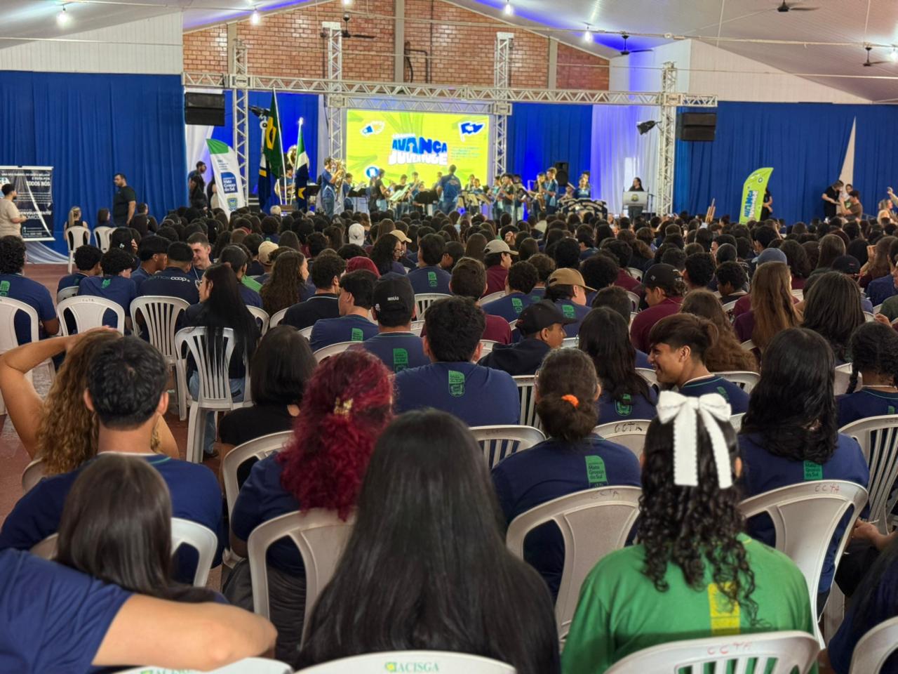 Vista panorâmica de um grande auditório lotado de jovens sentados em cadeiras brancas. Ao fundo, um palco com iluminação azul, bandeiras e um telão exibindo a logomarca "Avança Juventude". Uma banda marcial se apresenta no palco.