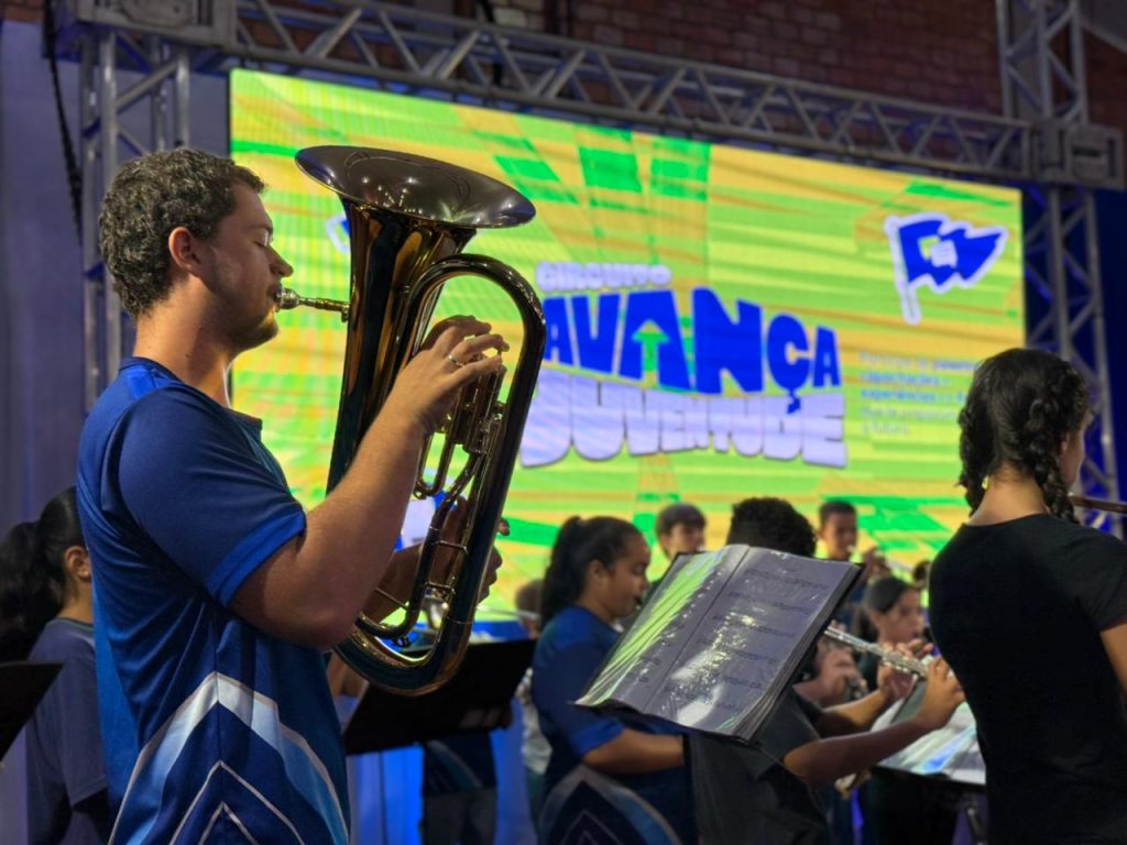 Close em um jovem integrante da banda tocando um bombardino (instrumento de sopro dourado). Ao fundo, o telão brilhante do evento e outros músicos da orquestra em desfoque.