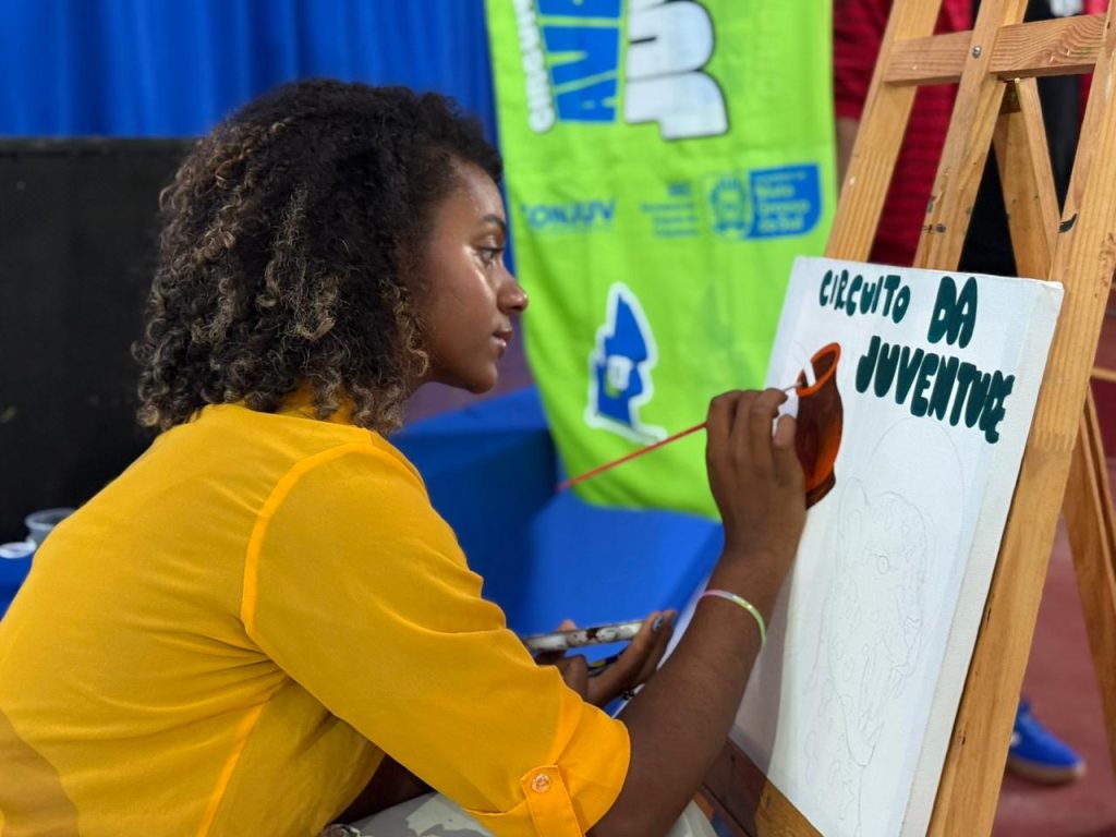Uma jovem de perfil, com cabelos cacheados e camisa amarela, pinta cuidadosamente uma tela em um cavalete de madeira. Na tela, lê-se "Circuito da Juventude" acima de uma pintura em progresso.