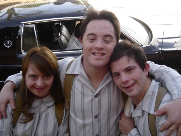 Fotografia colorida em plano médio de três jovens adultos com síndrome de Down, sorrindo abraçados. À esquerda, uma mulher de cabelos castanhos; ao centro, um homem mais alto com os braços sobre os ombros dos colegas; e à direita, outro homem encostado no colega central. Todos vestem camisas de botões bege com listras finas verticais e usam mochilas. Ao fundo, vê-se a lateral de um carro antigo preto e luz solar filtrada pelas árvores.