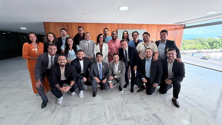 Fotografia em grupo de aproximadamente 20 pessoas em um saguão de piso claro e brilhante. O grupo é diverso e está organizado em duas fileiras: alguns agachados à frente e outros em pé atrás. A maioria veste trajes casuais e formais, como blazers e camisas sociais. Ao fundo, há uma parede revestida de madeira e, à direita, uma grande janela de vidro que mostra uma vista externa arborizada. Todos sorriem para a foto em um clima de confraternização.