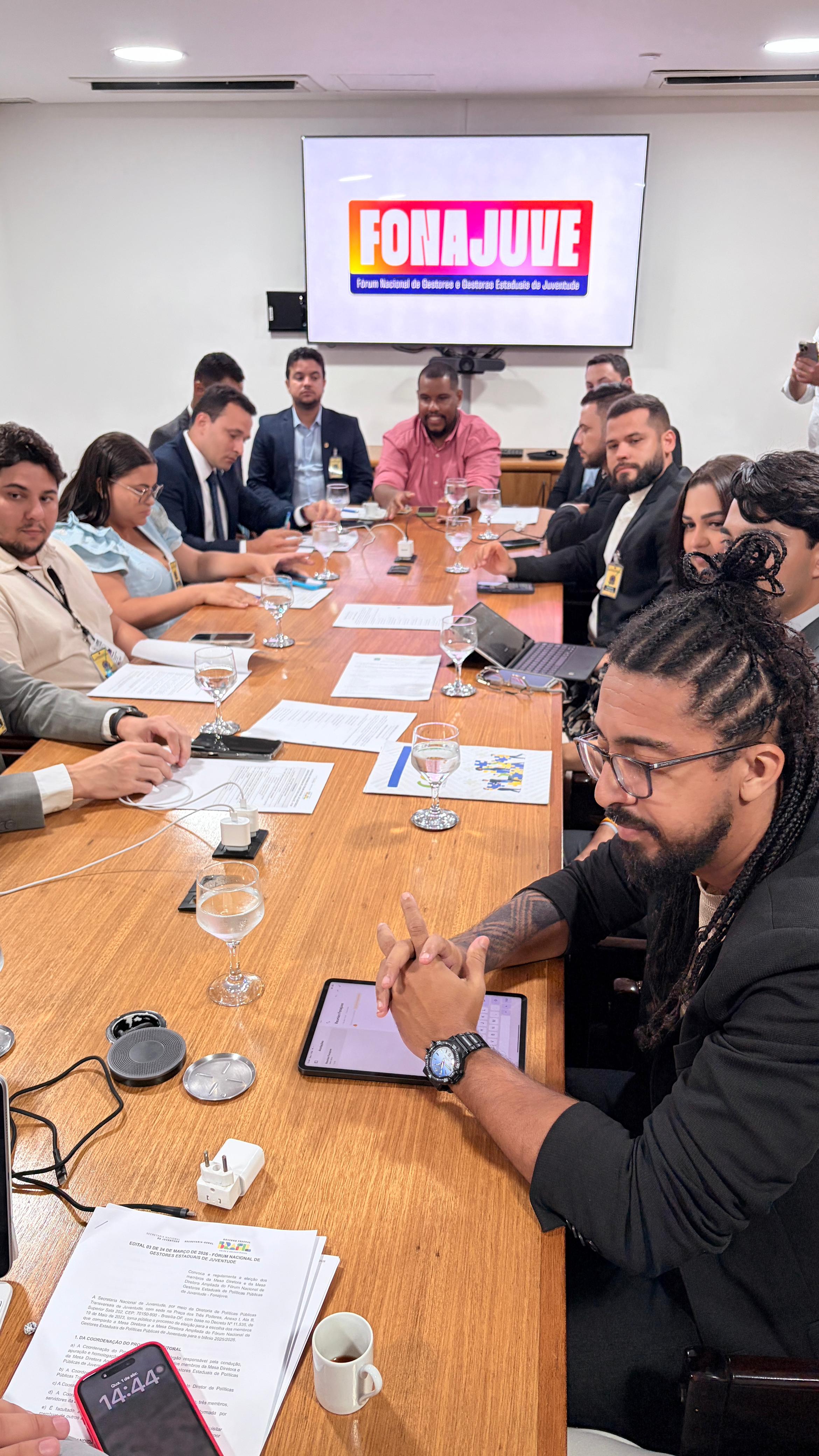 Fotografia em plano médio de uma reunião em torno de uma grande mesa de madeira clara. Diversas pessoas, homens e mulheres, estão sentadas discutindo documentos. Em primeiro plano, à direita, um homem negro com cabelos em tranças (dreadlocks) e barba usa óculos e um blazer preto, olhando atentamente para um tablet sobre a mesa. Ao fundo, uma TV fixada na parede exibe a logomarca colorida do "FONAJUVE - Fórum Nacional de Gestoras e Gestores Estaduais de Juventude". Sobre a mesa, há copos com água, papéis e dispositivos eletrônicos.