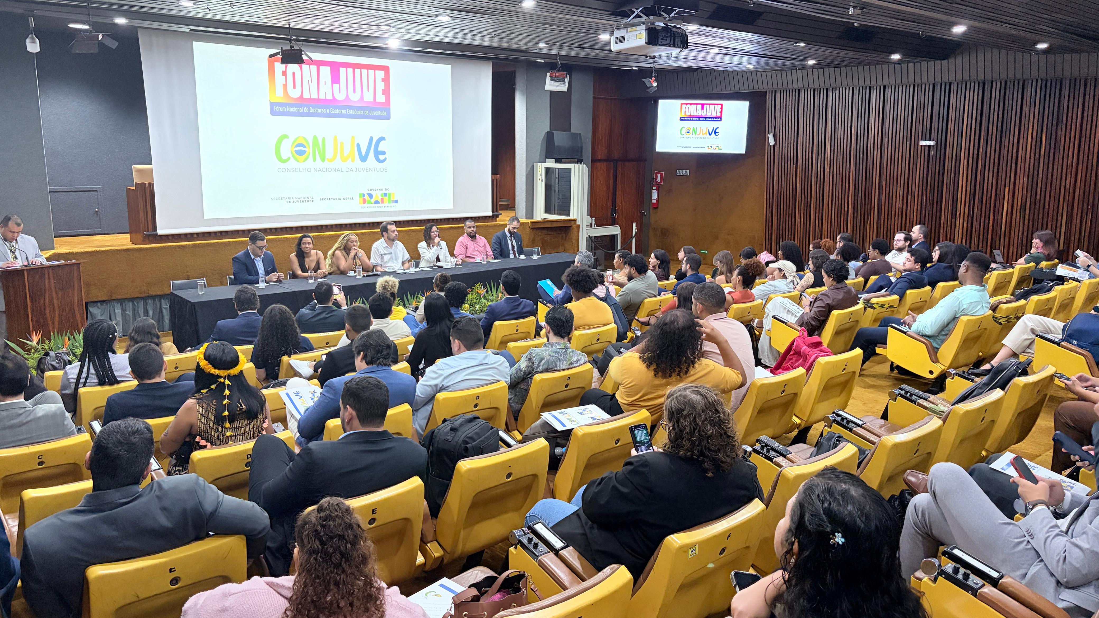 Fotografia em ângulo aberto de um auditório lotado durante um evento. No palco, uma mesa diretiva composta por sete pessoas está posicionada à frente de um grande telão branco que exibe as logomarcas do "FONAJUVE" e do "CONJUVE". O auditório possui poltronas amarelas ocupadas por diversos participantes de costas para a câmera. O teto é inclinado com forro de madeira e as paredes laterais também possuem revestimento amadeirado. No lado esquerdo, um homem fala em um púlpito de madeira.