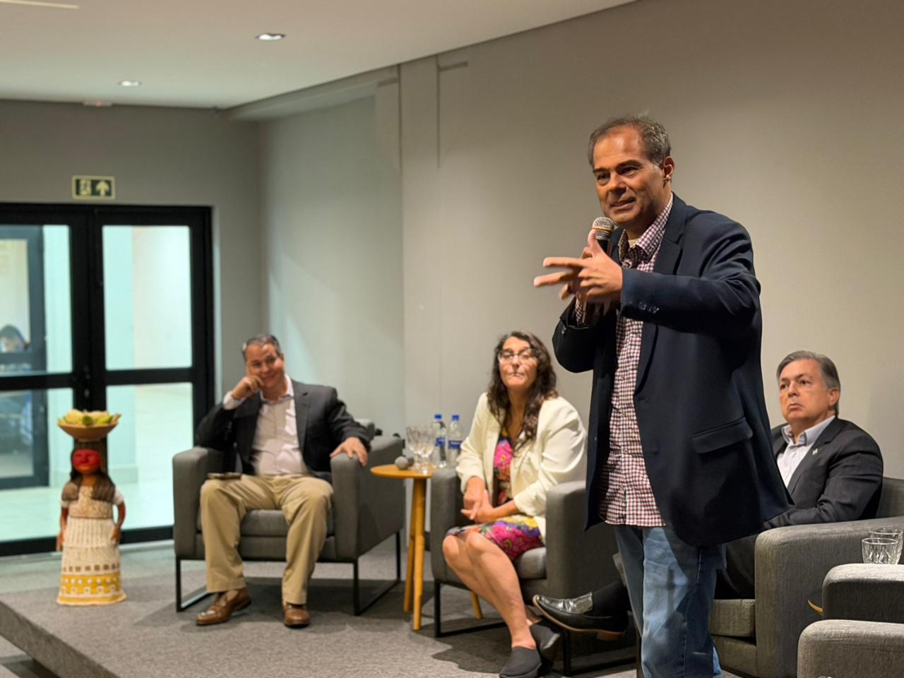 Fotografia em plano médio de um homem em pé, à direita, falando ao público em um auditório. Ele é pardo, tem cabelos curtos e grisalhos, veste um paletó azul-marinho sobre uma camisa xadrez miúda e calça jeans. Ele segura um microfone com a mão direita e gesticula com a esquerda. Ao fundo, três pessoas estão sentadas em poltronas cinzas: um homem à esquerda sorrindo, uma mulher ao centro com blazer branco e um homem à direita com expressão séria. No canto esquerdo, há uma estátua artesanal de uma figura indígena carregando uma cesta na cabeça. O ambiente tem iluminação suave e paredes em tons de cinza.