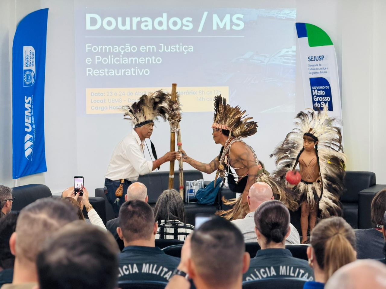 Momento de interação no palco entre dois indígenas que seguram juntos um bastão de madeira (borduna). A criança indígena observa ao lado. A plateia, composta majoritariamente por policiais, observa a cena.