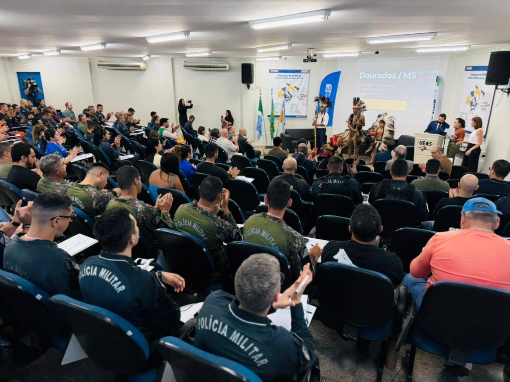 Visão panorâmica do auditório lotado. Nas primeiras fileiras, vários policiais militares fardados assistem à apresentação dos indígenas no palco. O ambiente é amplo, com cadeiras azuis e iluminação clara.