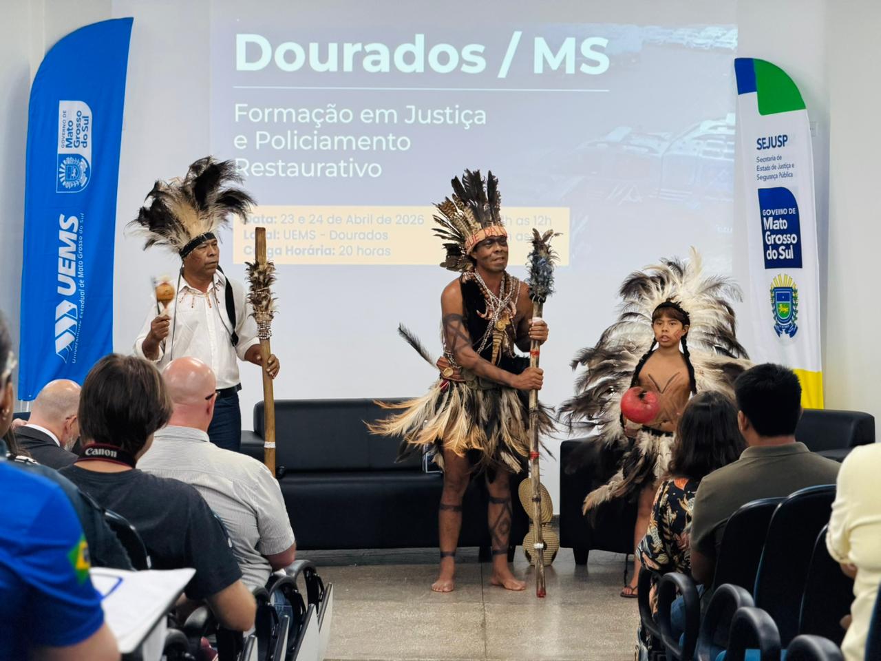 Três indígenas (dois adultos e uma criança) em pé à frente de um telão. Eles vestem trajes tradicionais com grandes cocares de penas escuras e claras, saias de fibras naturais e pinturas corporais. Os adultos seguram bordunas e maracás. Ao fundo, um telão anuncia o evento em Dourados/MS.
