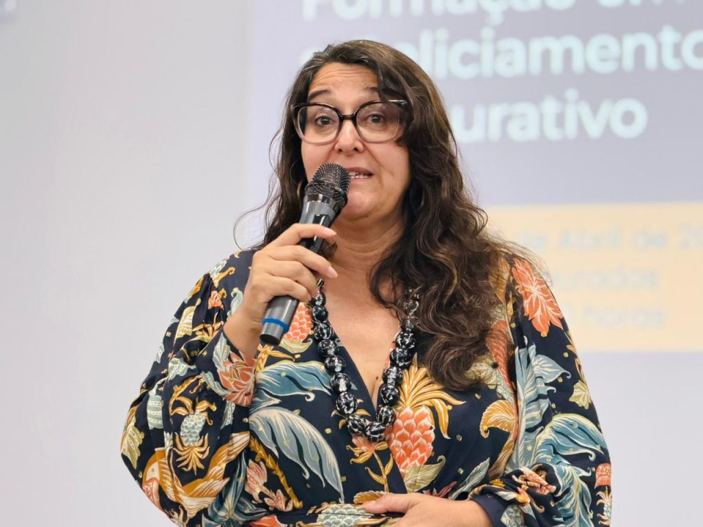 Close de uma palestrante com óculos e cabelos castanhos ondulados, usando um vestido estampado com motivos de abacaxi e um colar de contas pretas. Ela fala ao microfone.