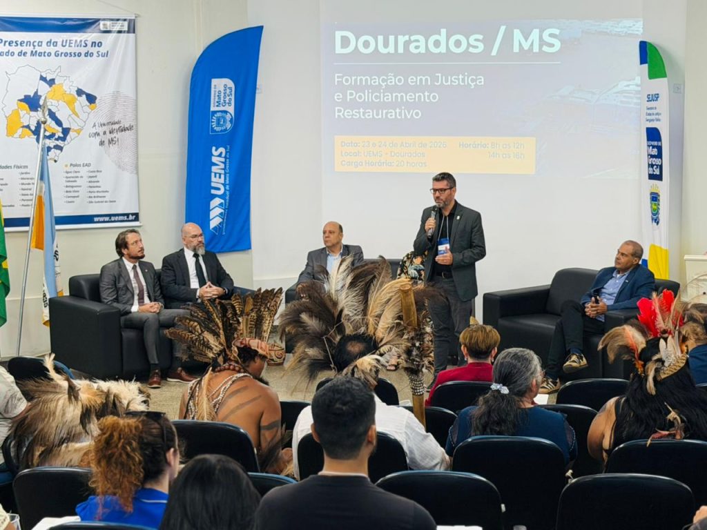 Um homem de paletó cinza fala ao microfone em um painel de discussão. Ele está em pé à frente de um sofá onde outros quatro palestrantes estão sentados. O público é visto de costas em primeiro plano.