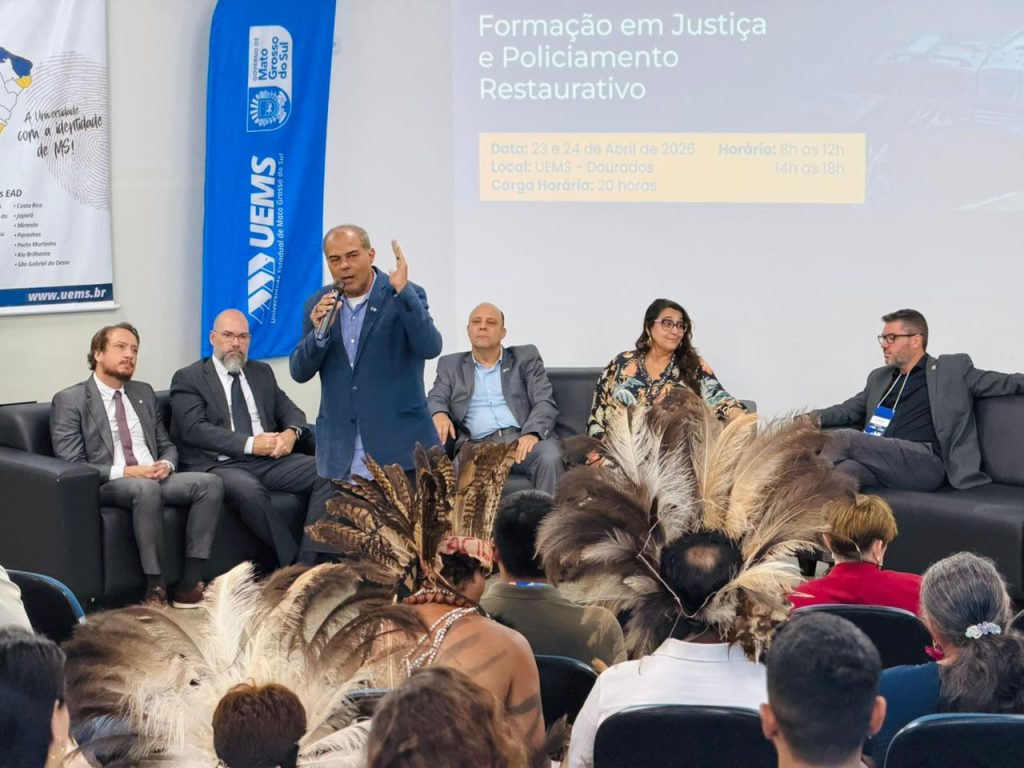 Um homem de blazer azul faz um gesto com a mão enquanto fala ao microfone. Ele compõe a mesa de palestrantes ao lado de outros homens e da mulher de vestido estampado.