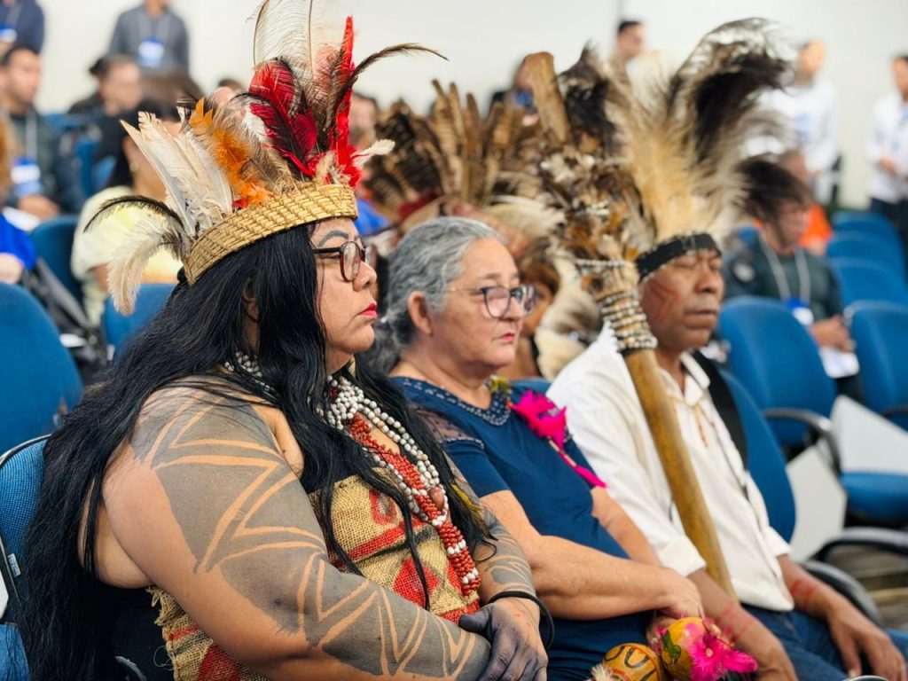 Close de uma mulher indígena sentada em um auditório, usando um cocar de penas vermelhas e amarelas, colares de contas e pinturas geométricas nos braços. Ao lado dela, outras pessoas assistem atentamente à palestra.